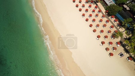 Foto de Vista superior de un club de playa con piscina, tumbonas y sombrillas rojas en la playa de arena blanca. Playa de arena blanca, corales, agua turquesa y un lujoso club en la costa de Bali. - Imagen libre de derechos