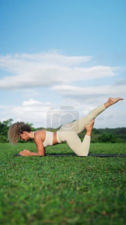 Foto de Una joven atlética latina atractiva está haciendo yoga al aire libre sobre una alfombra en el contexto de un bosque verde. Un entrenador de yoga realiza asana en el parque al amanecer. Prácticas de bienestar sobre lo natural. - Imagen libre de derechos