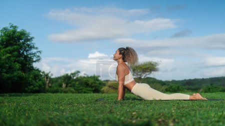 Foto de La pancarta de una joven atlética latina atractiva está haciendo yoga al aire libre sobre una alfombra en el contexto de un bosque verde y respirando profundamente. Un entrenador de yoga realiza asana en el parque al amanecer - Imagen libre de derechos