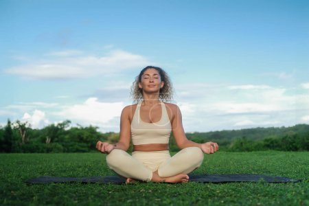 Photo for A young Latino woman with curly hair sits in the lotus position on a yoga mat and meditates against the backdrop of a green park at dawn. Concentration of breath and mind. Mindfulness practice. - Royalty Free Image