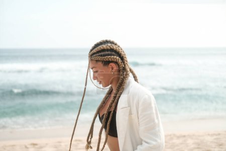 Foto de Retrato de una joven latina con trenzas en la cabeza. Camina por la playa cerca del océano con una chaqueta y un traje. Concepto de autoconfianza y masculinidad. Estudio de trenzas. - Imagen libre de derechos