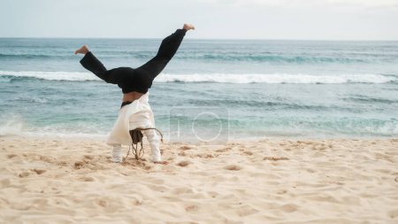 Téléchargez les photos : Vue arrière d'une jeune fille avec des tresses faisant un handstand sur la plage près de l'océan. Loisirs amusants et actifs sur la côte, sports de plein air. Mode de vie actif. Une femme fait une roue de chariot. - en image libre de droit