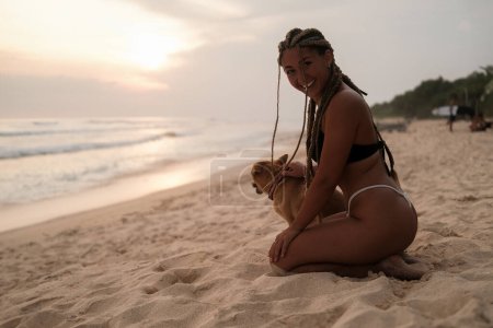 Foto de Joven latina sexy en bikini con trenzas en la cabeza. Sentado en la playa al atardecer con su perro y acariciándolo. Relajación y paseos por la costa oceánica con tu mascota. - Imagen libre de derechos