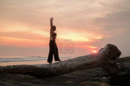Foto de Silueta de una joven con trenzas al atardecer en la playa, que camina a lo largo de un tronco caído y extiende los brazos hacia arriba. Sensual danza junto al océano durante la hora dorada al atardecer. - Imagen libre de derechos