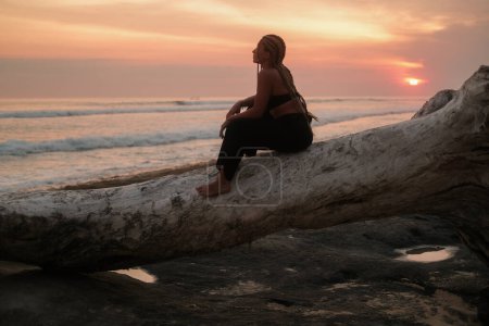 Téléchargez les photos : Silhouette d'une jeune femme avec des tresses au coucher du soleil sur la plage, posant sur une bûche tombée. Danse sensuelle sur l'océan à l'heure dorée du coucher du soleil. Présentation d'un corps maigre athlétique en vacances. - en image libre de droit