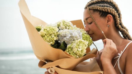 Photo for Portrait of a young woman with braids smelling a bouquet of hydrangea flowers on the beach near the ocean. A gift for your girlfriend on Valentines Day and International Womens Day. - Royalty Free Image