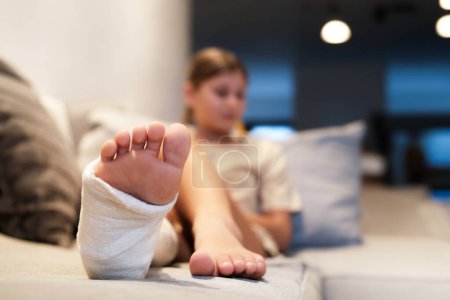 A teenage girl with a cast on her leg sits in the living room on the sofa. A teenager has a broken leg with crutches in the background. Recovery after injury.