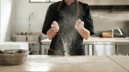 Téléchargez les photos : Gros plan sur les mains d'une pâtissière dont les mains sont couvertes de farine et elle applaudit ses mains sur le fond d'un tablier noir créant un nuage blanc de farine Préparation de la pâte dans une boulangerie - en image libre de droit