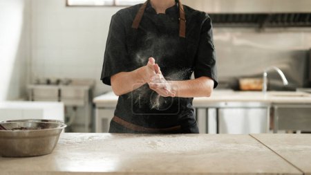 Großaufnahme der Hände einer Konditorin, deren Hände mit Mehl bedeckt sind, und sie klatscht in die Hände vor dem Hintergrund einer schwarzen Schürze, die eine weiße Wolke aus Mehl schafft.