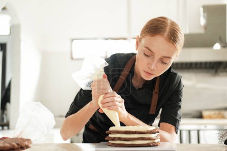 Foto de Retrato de una joven pastelera pelirroja aplicando crema de mantequilla a una capa de pastel de chocolate usando una bolsa de pastelería. El proceso de preparación de un pastel en una pastelería en la cocina profesional. - Imagen libre de derechos