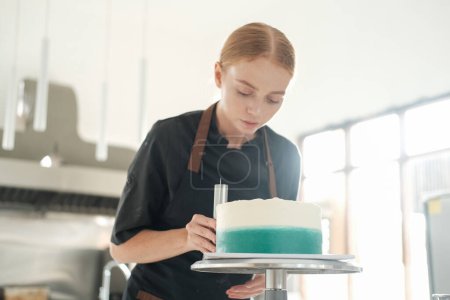 Téléchargez les photos : Une jeune pâtissière rousse distribue de la crème turquoise sur un gâteau à l'aide d'une spatule sur un support rotatif. Le processus de préparation du dessert dans une confiserie. - en image libre de droit