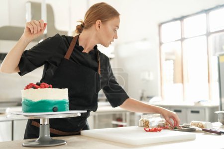 Foto de Retrato cercano de una joven y encantadora chef de pastelería pelirroja que se concentra decorando un pastel con bayas frescas. El proceso de trabajo en una confitería profesional. Confitería. - Imagen libre de derechos