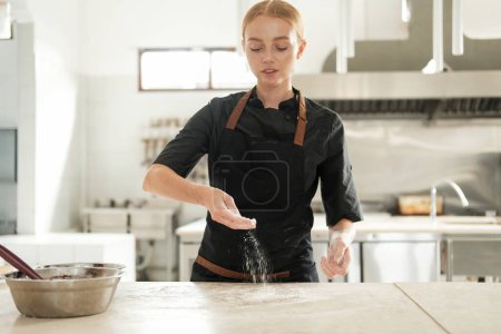 Téléchargez les photos : Plan moyen des mains d'une femme qui saupoudre la farine sur la surface de la table au ralenti sur le fond d'un tablier noir. Le processus de préparation de la pâte sur le plancher de production dans une boulangerie. - en image libre de droit