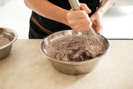 Téléchargez les photos : Gros plan des mains d'une jeune pâtissière dans un tablier, qui pétrit la farine et la poudre de cacao dans un bol pour préparer des couches de gâteau au chocolat. Le processus de préparation de la pâte. - en image libre de droit