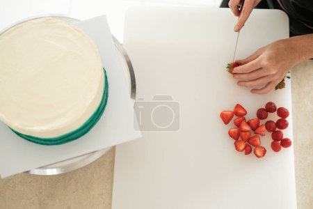 Téléchargez les photos : Vue de dessus du plan de travail d'une table dans une pâtisserie. Gros plan des mains d'une jeune pâtissière qui découpe des baies fraîches pour décorer des gâteaux sur une planche à découper blanche. - en image libre de droit