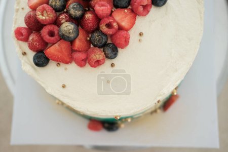 Foto de Vista superior de un pastel cubierto de turquesa y crema blanca, decorado con bayas frescas. Se asienta en un stand giratorio en el área de producción de una pastelería. Hermosa pancarta de pastel de cumpleaños - Imagen libre de derechos