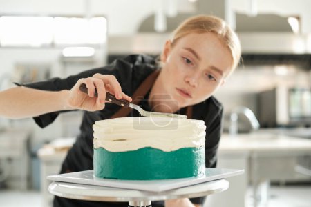 Photo for Portrait of a young red haired female pastry chef who looks at the cake with concentration and distributes the cream in an even layer over its surface. The process of preparing in a production kitchen - Royalty Free Image