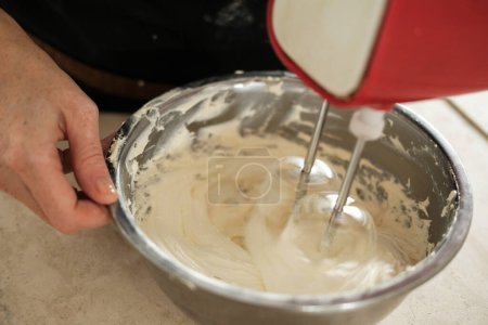 Photo for A close-up of the hands of a young woman pastry chef who is whipping butter cream for a cake using an electric mixer in a deep iron bowl. The process of preparation in production in a confectionery. - Royalty Free Image