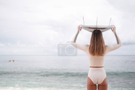 Téléchargez les photos : Vue arrière bannière d'une jeune femme sexy debout sur une plage près de l'océan portant un bikini de surf blanc et tenant une planche de surf sur la tête. Bonne jeune surfeuse sportive. Allons surfer. - en image libre de droit