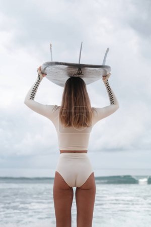 Foto de Vista trasera de una joven rubia parada en la playa en un traje de baño de surf blanco y sosteniendo una tabla de surf en su cabeza mirando al océano. Feliz joven surfista en bikini. Vamos a surfear. - Imagen libre de derechos