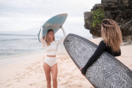 Photo for Banner with two diverse young women in white and black surf bikinis talking while standing on the beach with their surfboards. Womens surf community. Friendship in extreme water sports. - Royalty Free Image