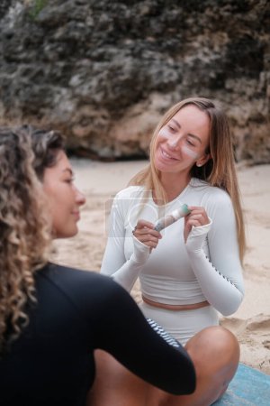 Téléchargez les photos : Une jeune femme blonde tenant de la crème solaire zinc pour surfer, regardant la caméra et souriant. Son amie est assise à côté d'elle sur une planche de surf. Protection de la peau pour les surfeurs. Crème imperméable. - en image libre de droit