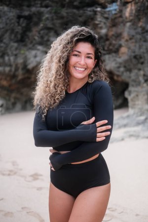 Photo for Young attractive Latina woman with curly hair stands on the beach wearing a black long sleeve surf swimsuit and smile. Advertising for womens surf wear. Comfortable sportswear. - Royalty Free Image