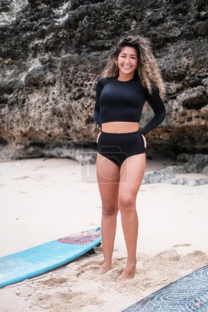 Foto de Una joven latina con el pelo rizado en un traje de baño de surf negro está en la playa cerca del océano y hace un calentamiento con una tabla de surf acostada a su lado. La mujer sonríe y disfruta del surf. - Imagen libre de derechos