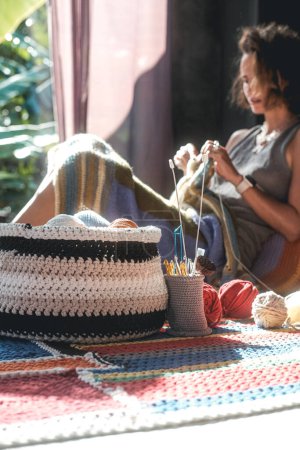 Téléchargez les photos : Une femme est assise sur le sol et tricote une couverture multicolore à partir de fils de laine au premier plan est un panier avec des boules de laine en arrière-plan est une fenêtre derrière laquelle il ya de la verdure tropicale - en image libre de droit
