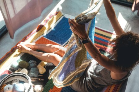 Téléchargez les photos : Une jeune femme tricote une couverture multicolore et douce à partir de fils de laine. Elle l'a soulevée pour évaluer la qualité de son travail. Elle s'assoit par terre dans sa chambre, un passe-temps tranquille. - en image libre de droit