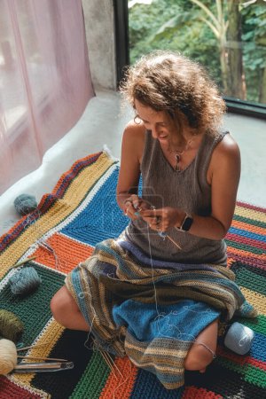 Téléchargez les photos : Une femme s'assoit par terre dans la chambre et tricote une couverture à partir de fils de laine. Le fond de la fenêtre est la verdure tropicale. A côté d'elle se trouve un panier avec des boules de laine et des outils à tricoter. - en image libre de droit