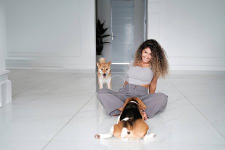 Téléchargez les photos : Bannière avec une jeune femme aux cheveux bouclés assise sur le sol en vêtements décontractés à côté de ses deux chiens. L'un est un chien Shiba Inu et l'autre est un chien Beagle. Ils jouent sur le sol. - en image libre de droit