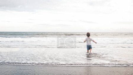 Téléchargez les photos : Un jeune garçon heureux court joyeusement dans l'eau de l'océan sur la plage, sous le ciel bleu et les nuages pelucheux. Les vagues liquides et fluides éclaboussent autour de lui alors que le vent souffle doucement. Fête des mères. - en image libre de droit