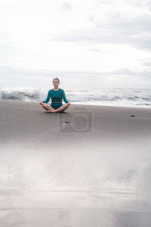 Téléchargez les photos : Une jeune femme en position lotus s'assoit sur une plage de sable noir et médite dans le contexte de l'océan avec de grandes vagues. Pratiquez la pleine conscience et la pureté de l'esprit. Concentration. Sports d'été. - en image libre de droit