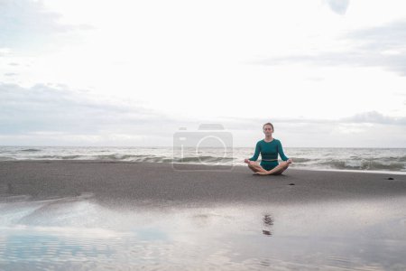 Téléchargez les photos : Bannière avec femme en position lotus assis sur une plage de sable noir et médite dans le contexte de l'océan avec de grandes vagues. Pratiquez la pleine conscience et la pureté de l'esprit. Concentration. Sports d'été. - en image libre de droit
