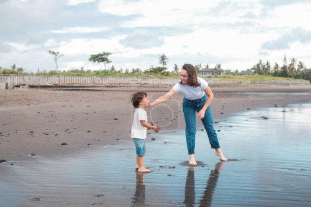 Photo for Mom and son are standing on a black beach near the ocean. They are dressed in casual clothes, the woman reaches out to the child and wipes his face. Taking care of your child on vacation. Mothers Day. - Royalty Free Image