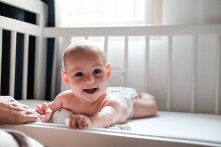 Téléchargez les photos : Portrait d'un bébé allongé dans un berceau dans une couche et riant, regardant la caméra, à côté de la main de sa mère. Joyeux et adorable nouveau-né avec un sourire sur le visage. La fête des mères. Sieste diurne - en image libre de droit