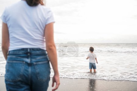 Foto de Mamá e hijo Los niños pequeños están caminando en la playa cerca del océano. En primer plano hay una silueta de una mujer en jeans viendo a su hijo chapotear en el agua. Día de las Madres. - Imagen libre de derechos
