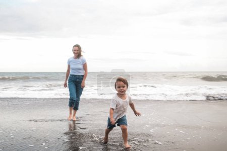 Téléchargez les photos : Maman et son fils Bébé courent le long de la plage de sable noir le long de l'océan et rient joyeusement. Ils jouent dans l'eau et passent du temps ensemble. Vacances d'été en famille. Fête des mères. - en image libre de droit