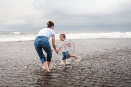 Foto de Mamá e hijo pequeño están corriendo a lo largo de la playa de arena negra a lo largo del océano y riendo felizmente. Juegan en el agua y pasan tiempo juntos. Vacaciones familiares de verano. Día de las Madres. - Imagen libre de derechos