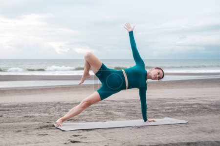 Téléchargez les photos : Une jeune femme en tenue de sport sur une plage de sable noir fait un yoga asana. Elle est concentrée et calme. Étirement et méditation au son des vagues. Prendre soin de votre corps. Style de vie estival. - en image libre de droit