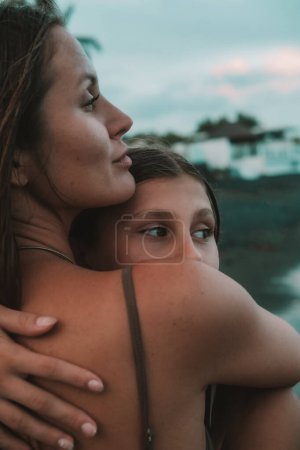 Foto de Un primer plano de una adolescente mirando por encima del hombro de su madre mientras la abraza en una playa de arena negra en la orilla del océano. Día de las Madres. Vacaciones con un niño en un país cálido. - Imagen libre de derechos