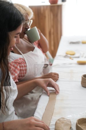 Vertical photo portrait of two diverse Caucasian and African American women who sitting in a pottery workshop and making mugs from clay. Creative activities for adults, hobbies with friend.