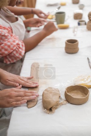 Vertical photo of the hands of two diverse Caucasian and African American women who sitting in a pottery workshop and making mugs from clay. Creative activities for adults, hobbies with friend.