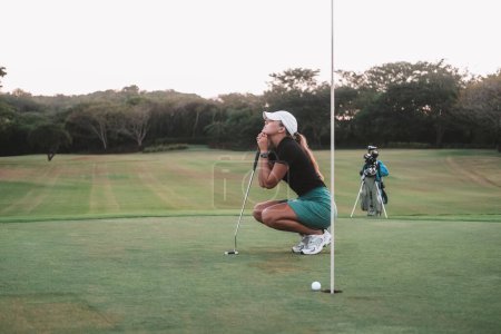 Una joven golfista principiante, se sienta en cuclillas en el campo de golf y sostiene un palo en sus manos. Ella está molesta porque se perdió el hoyo y perdió la competencia. Frustración de la derrota.