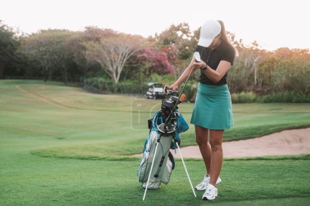 Banner with a young attractive woman who plays golf professionally. She stands on the green lawn next to her bag and selects a club to play. There is a golf cart in the distance in the background.