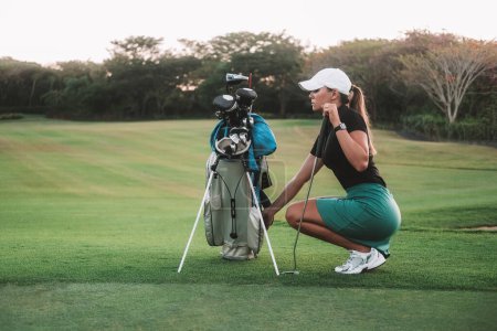 Banner with a young attractive woman who played golf professionally. She squats on the green lawn next to her bag and selects a club to play. Luxury summer sport.