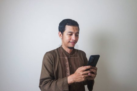 Cheerful young Asian Muslim man using a mobile phone and looking away isolated over white background