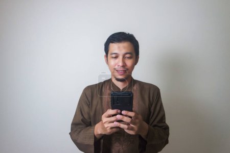 Fröhlicher junger asiatischer Muslim, der ein Mobiltelefon benutzt und isoliert vor weißem Hintergrund wegschaut