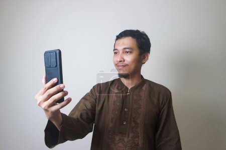 Cheerful young Asian Muslim man using a mobile phone and looking away isolated over white background